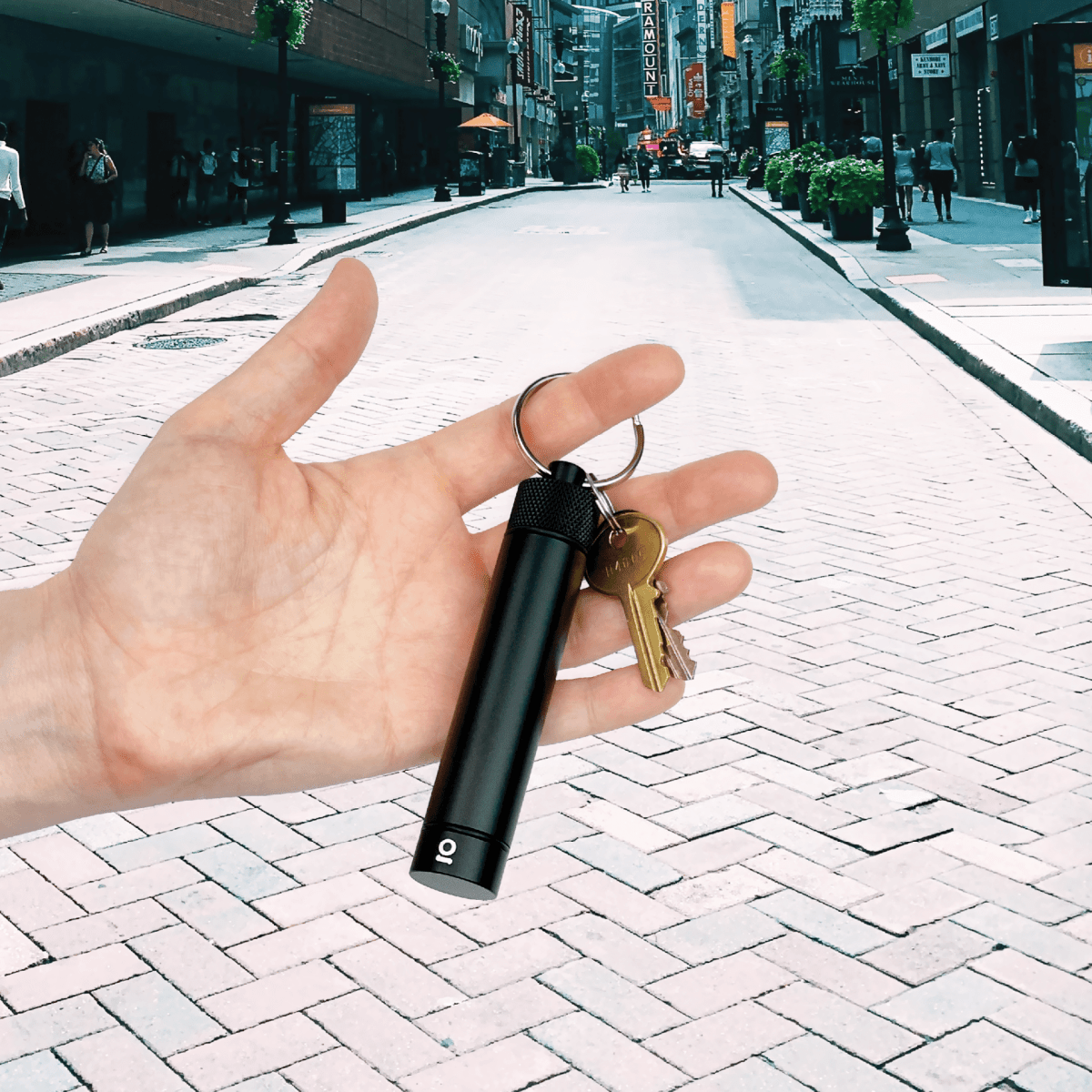 A hand displays the Aluminum Storage Keychain, a sleek, silver cylinder, against a blurred, sunlit city street background.