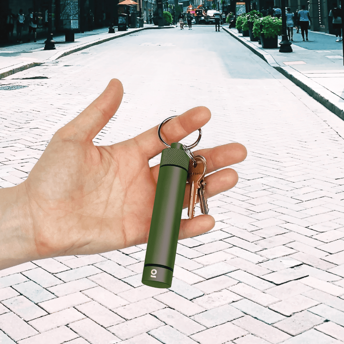 A hand holds the Aluminum Storage Keychain against a blurred background of a brick-paved street and foliage.