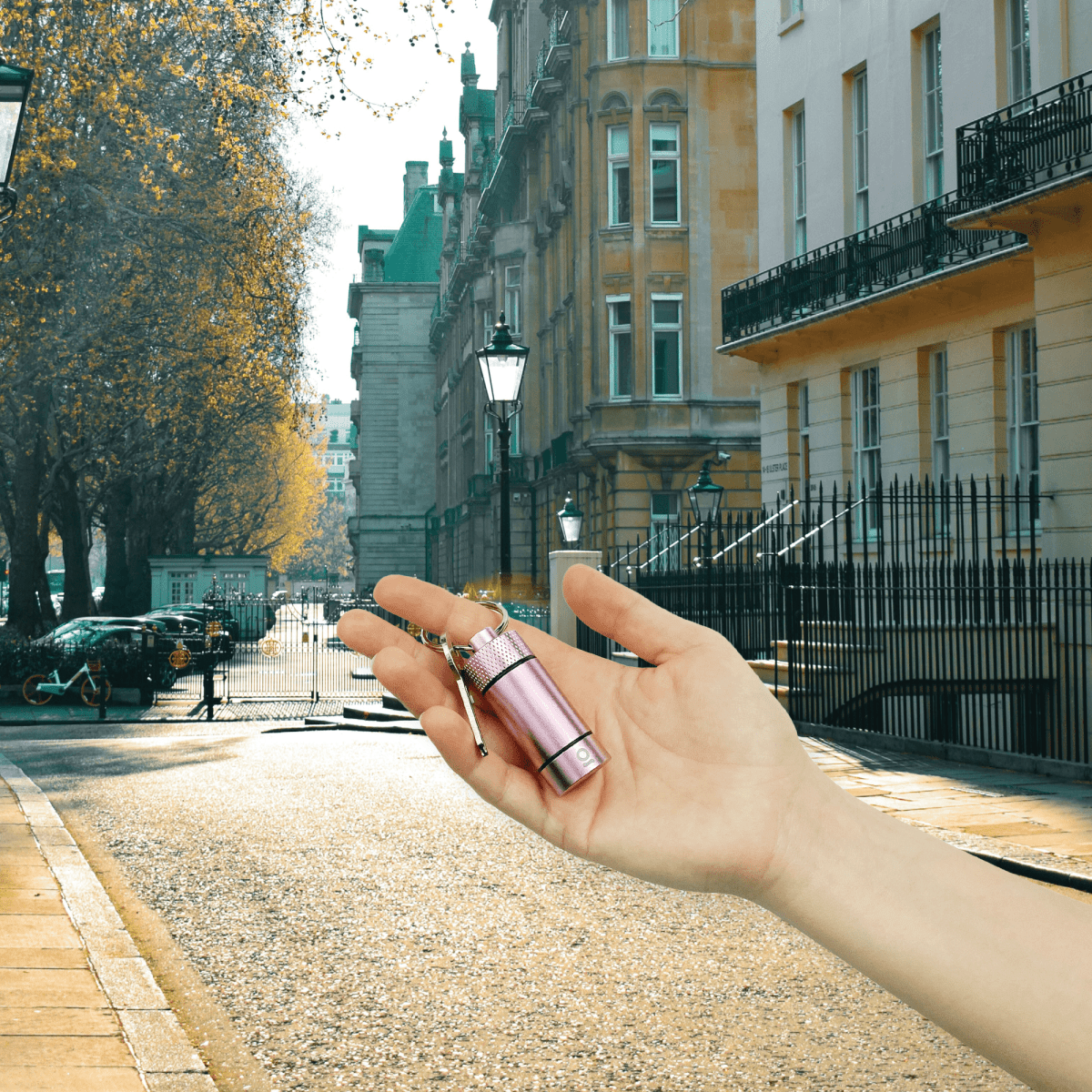 A hand holds the Small Storage Keychain against a blurred city street with classic architecture and a brick pathway.