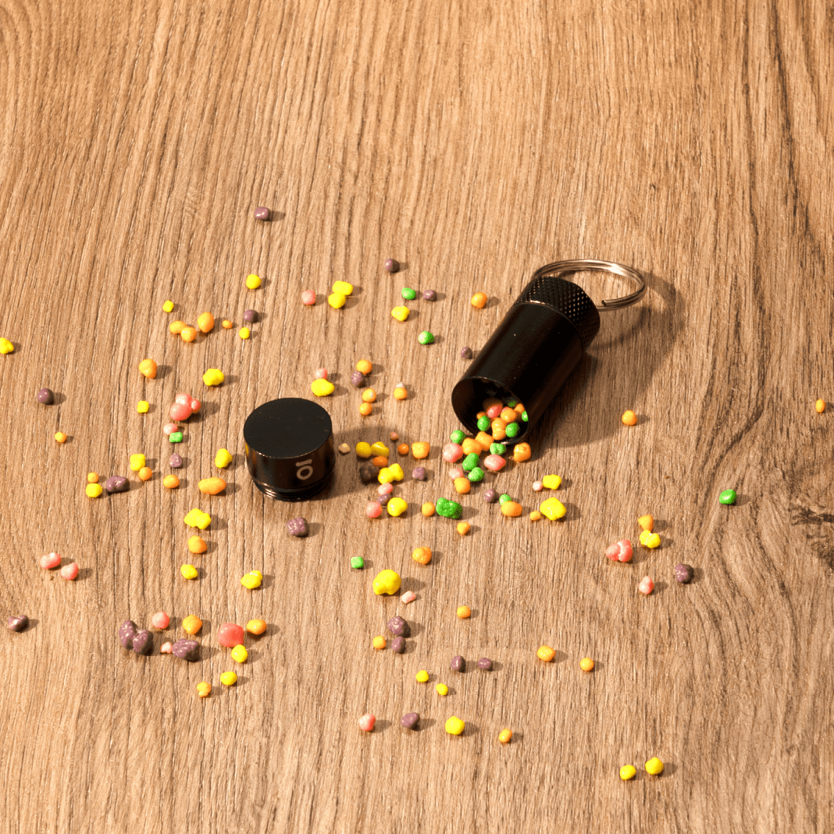Overhead view of the Small Storage Keychain, with colorful granules scattered around the dark-colored container on a wooden surface.