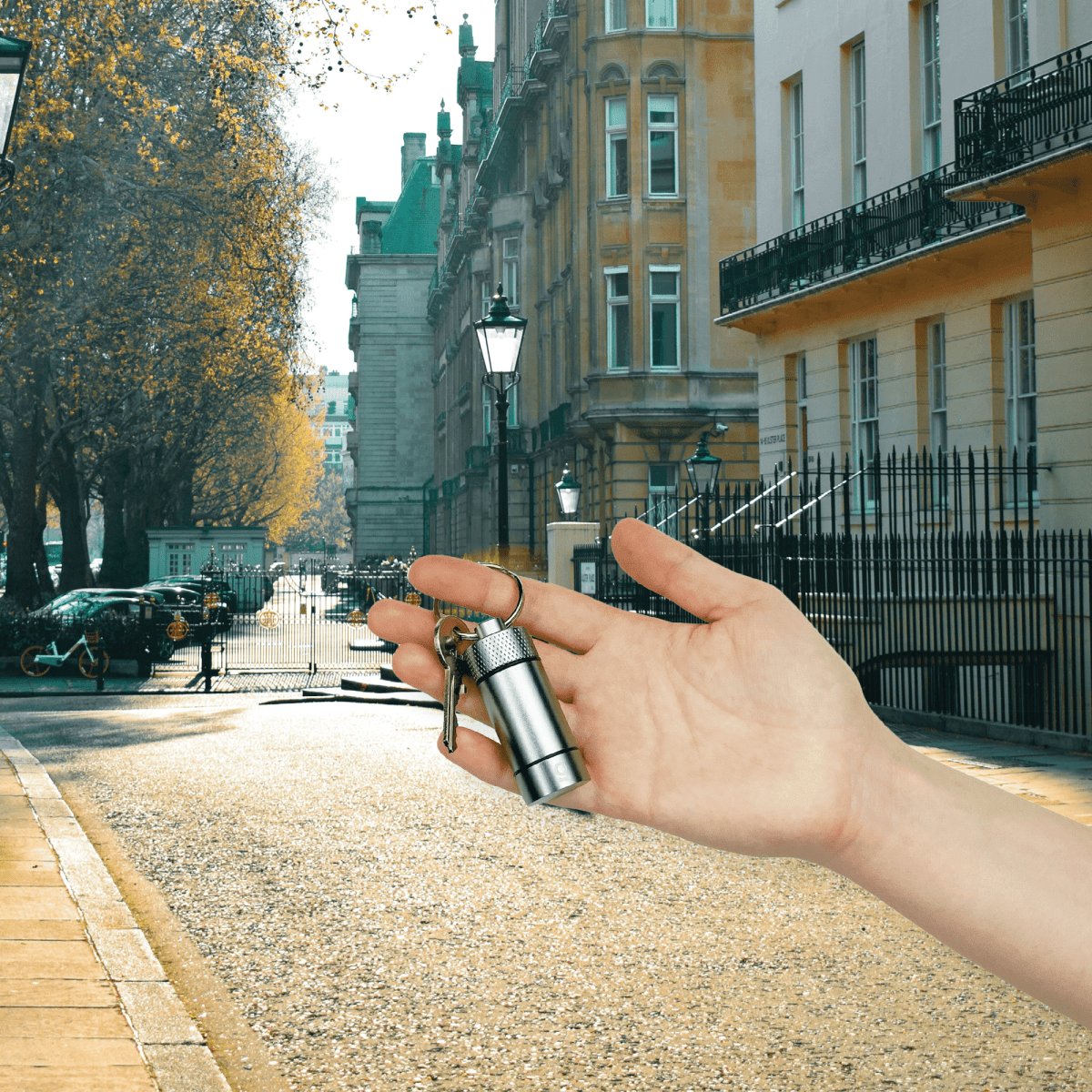 A hand holds the Small Storage Keychain against a backdrop of a European street with ornate buildings and a cobblestone path.
