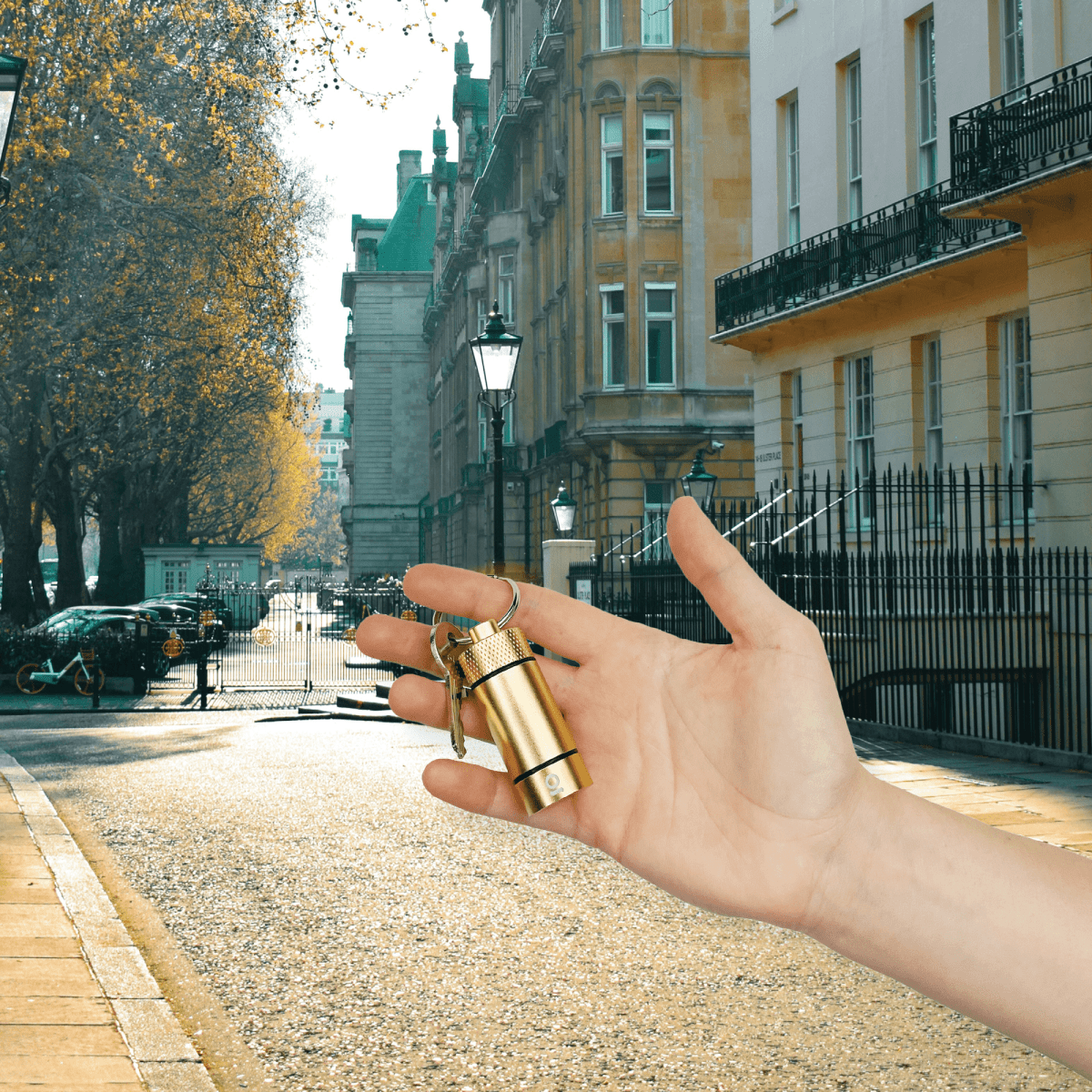 A hand holds the Small Storage Keychain against a blurred background of a cobblestone street and European-style buildings.