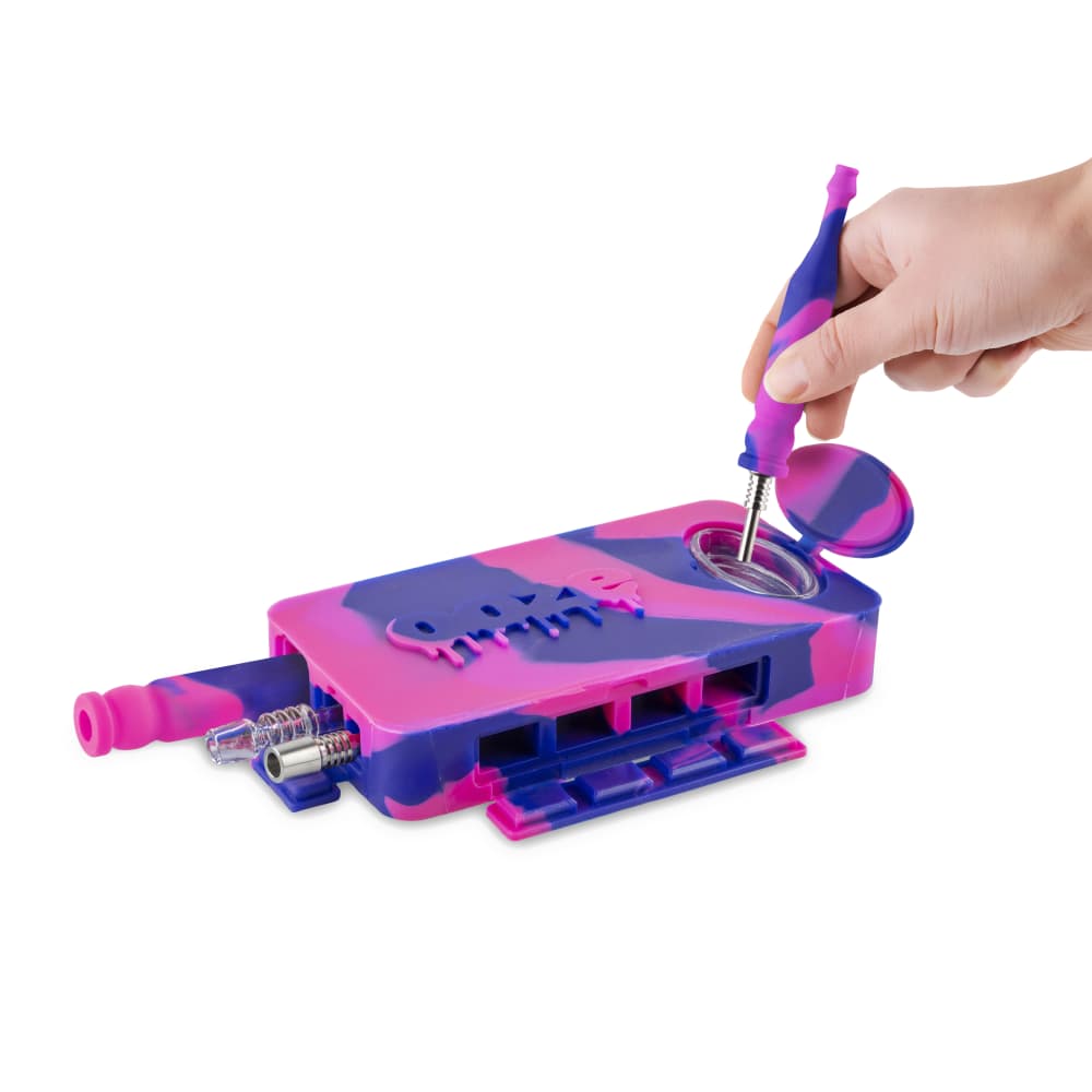 A hand holds a tool over the open Ooze Slugger Silicone Dugout, showcasing its dark interior against a bright white background.