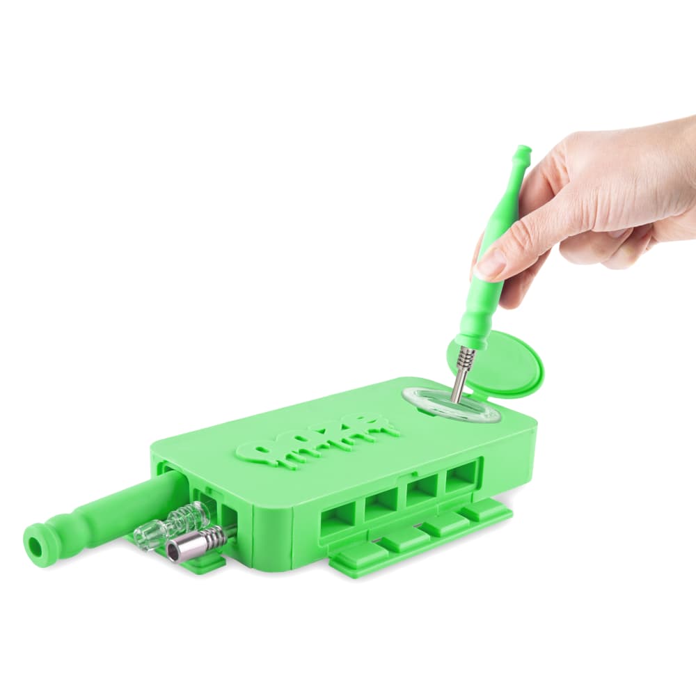 A hand holds a cleaning pick over the open Ooze Slugger Silicone Dugout, showing its interior detail against a white background.