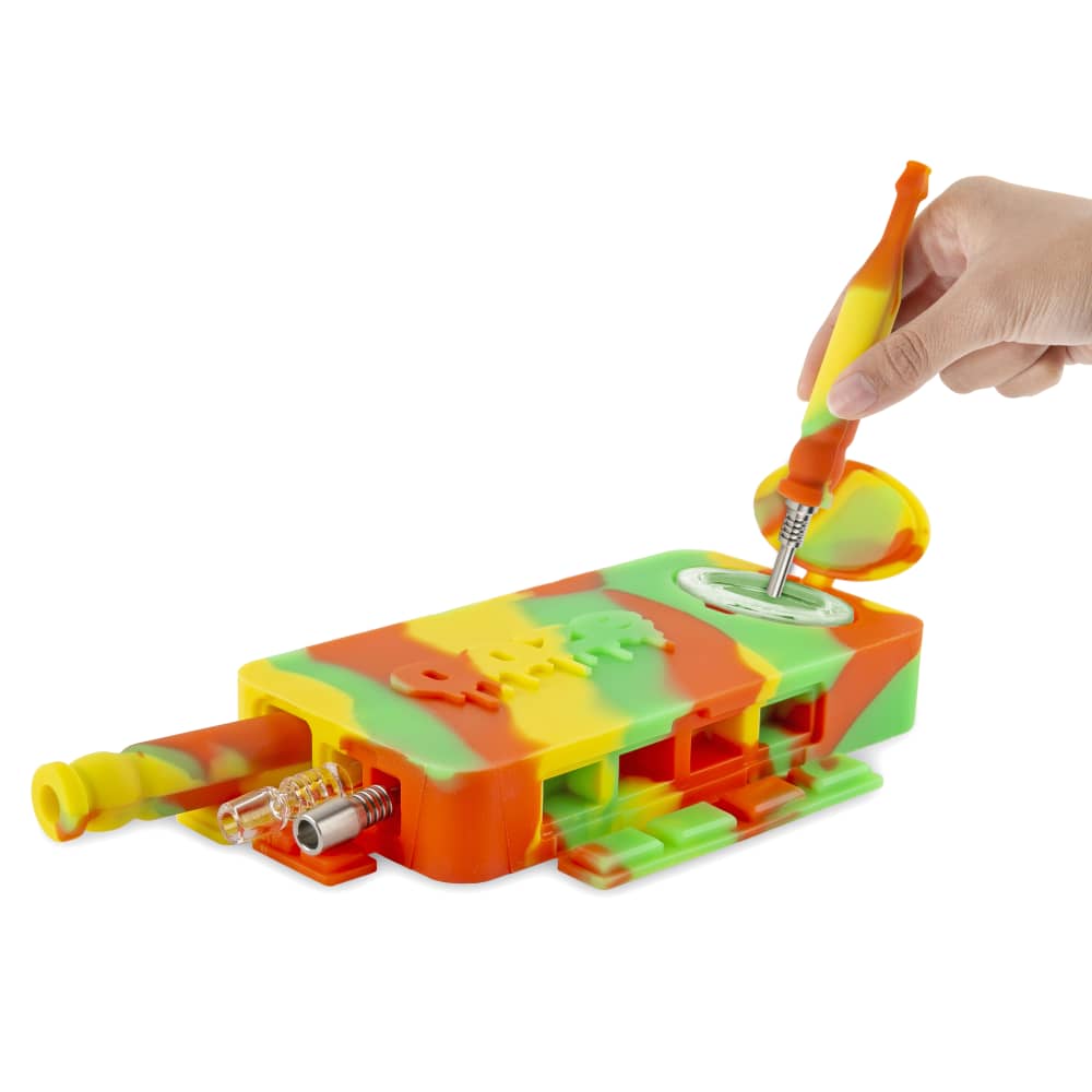 A hand holds a tool over the open compartment of the Ooze Slugger Silicone Dugout, revealing its interior against a plain white background.