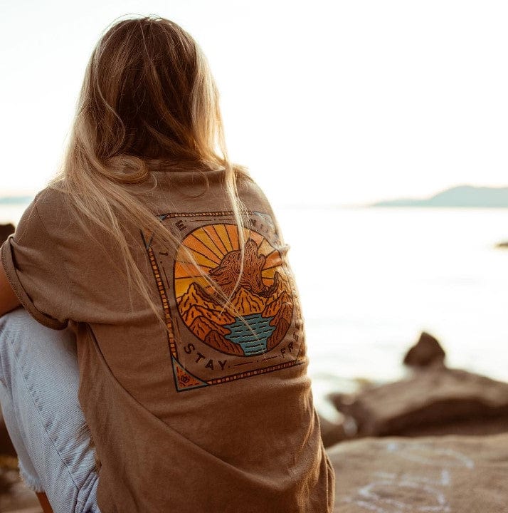 Rear three-quarter view of a person sitting by the water at sunset wearing a tan Prism Desert Dream'N Big Honeycomb Single Stack t‑shirt with a colorful mountain-and-sun graphic on the back.