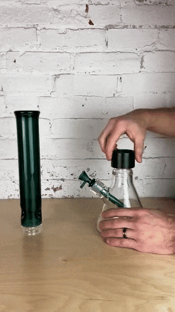 Hands assembling a Prism Percolated Beaker Double Stack on a wooden table, showing a clear beaker base and green cylindrical percolator against a white brick wall background.
