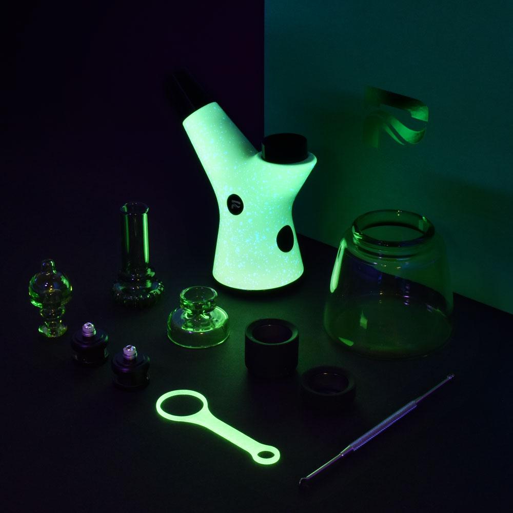 A moody studio shot displays the Pulsar RöK Electric Dab Rig - Luna Glow alongside small glass pieces on a dark surface.