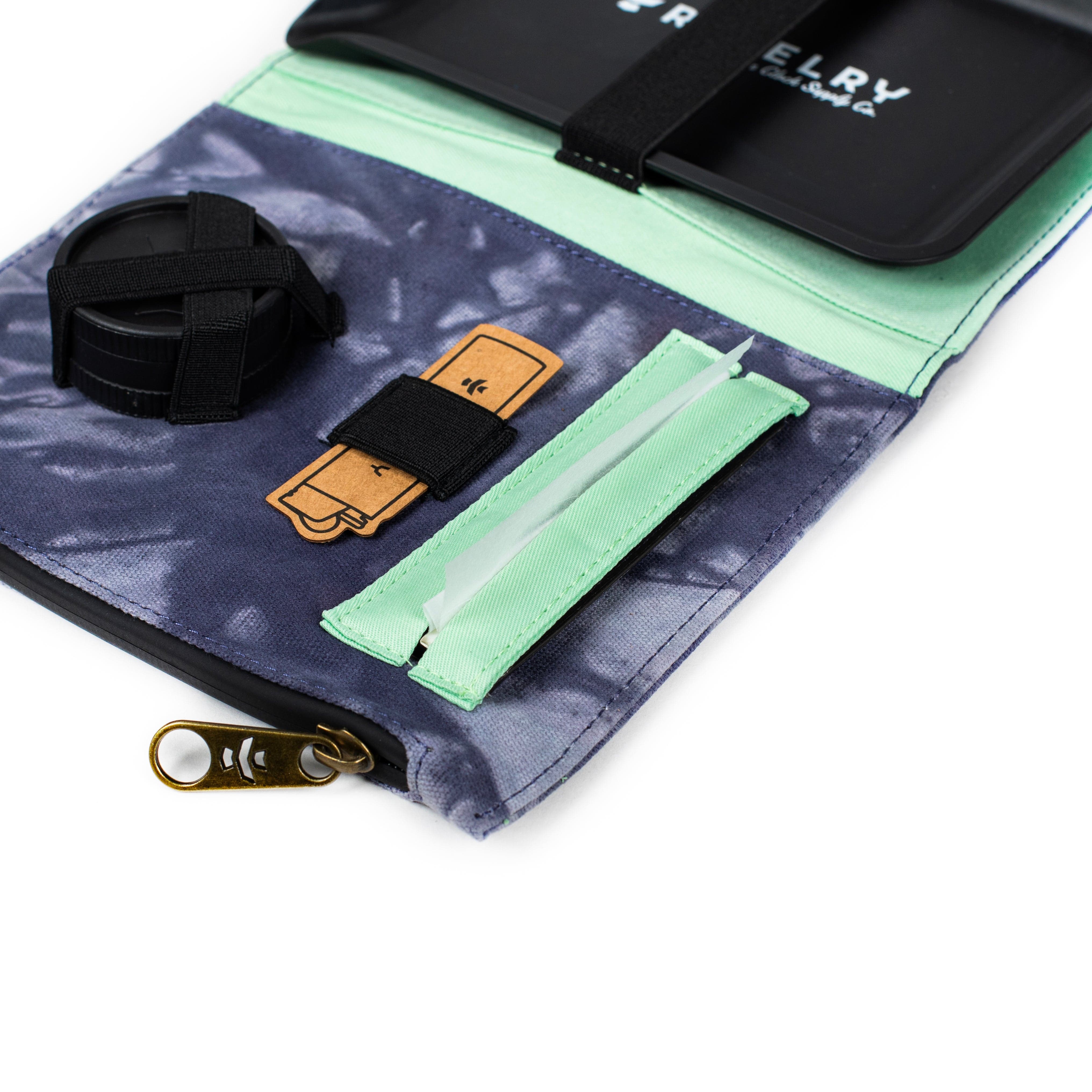 An angled view of The Rolling Kit - Smell Proof Kit open, revealing inner compartments and a zippered closure against a patterned fabric backdrop.